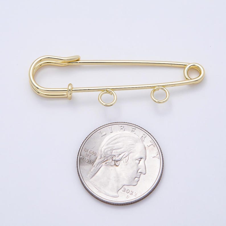 May include: A gold-toned metal safety pin featuring two small circular loops attached to the bottom bar. The safety pin is positioned above a standard United States quarter coin, which serves as a visual reference for its size and scale.