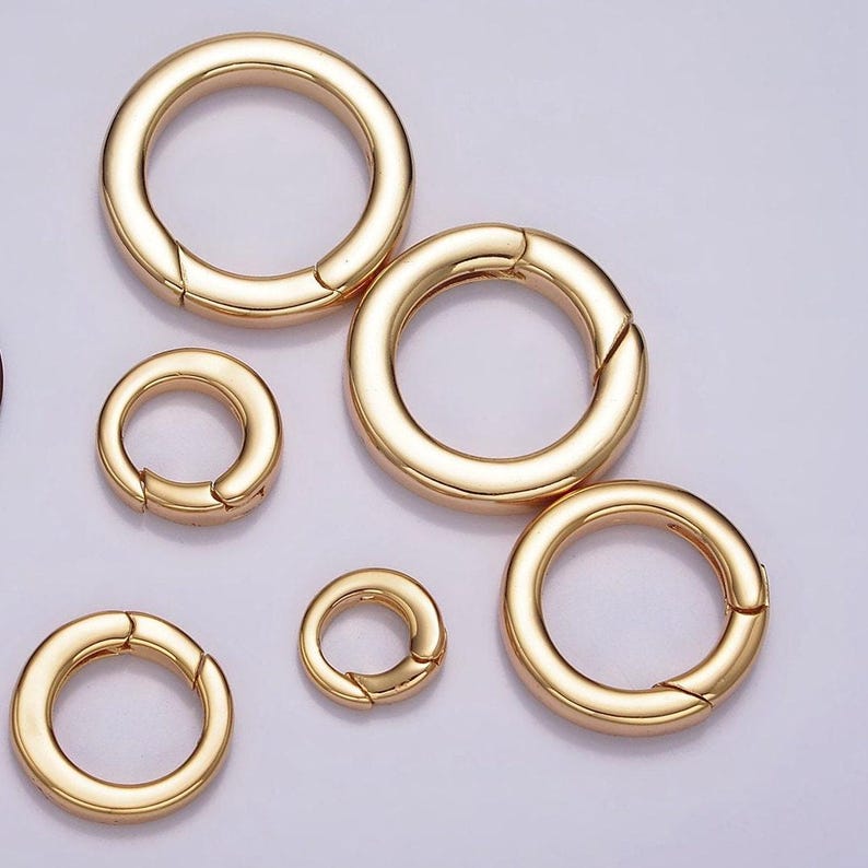 Pode incluir: Cinco an&eacute;is de metal dourado com fecho de mola. Os an&eacute;is s&atilde;o de tamanhos diferentes e est&atilde;o dispostos em semic&iacute;rculo sobre um fundo branco.