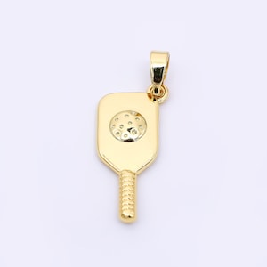 May include: A gold-colored pendant in the shape of a pickleball paddle. The paddle features a textured handle and a circular design with small indentations, resembling a pickleball. The pendant has a loop for a chain.