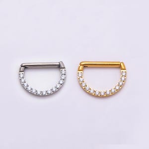 Puede incluir: Dos anillos de segmento en forma de D para piercing. Uno es de metal plateado y el otro de metal dorado. Ambos cuentan con una barra horizontal recta y una sección inferior curva incrustada con una hilera de pequeñas piedras claras.