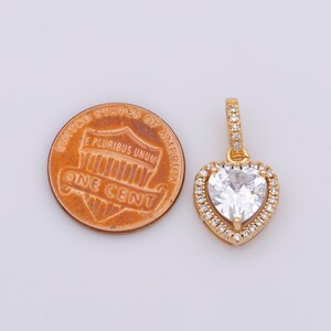 May include: A gold heart-shaped pendant featuring a large clear cubic zirconia stone surrounded by smaller clear cubic zirconia stones. The pendant is shown next to a UK penny for size comparison.