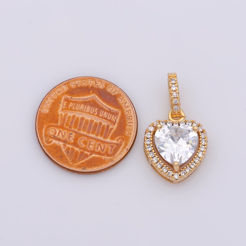 May include: A gold heart-shaped pendant featuring a large clear cubic zirconia stone surrounded by smaller clear cubic zirconia stones. The pendant is shown next to a UK penny for size comparison.