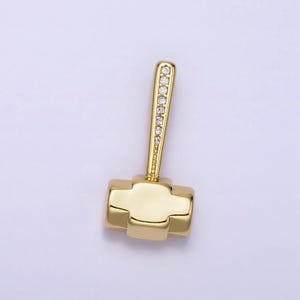 May include: A gold-colored hammer pendant with a handle adorned with small, clear gemstones. The hammerhead is rectangular with rounded edges. The pendant is set against a light background.
