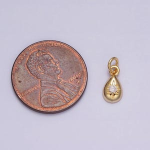 May include: A gold teardrop pendant with a small clear rhinestone in the center. The pendant is shown next to a US penny for size comparison.