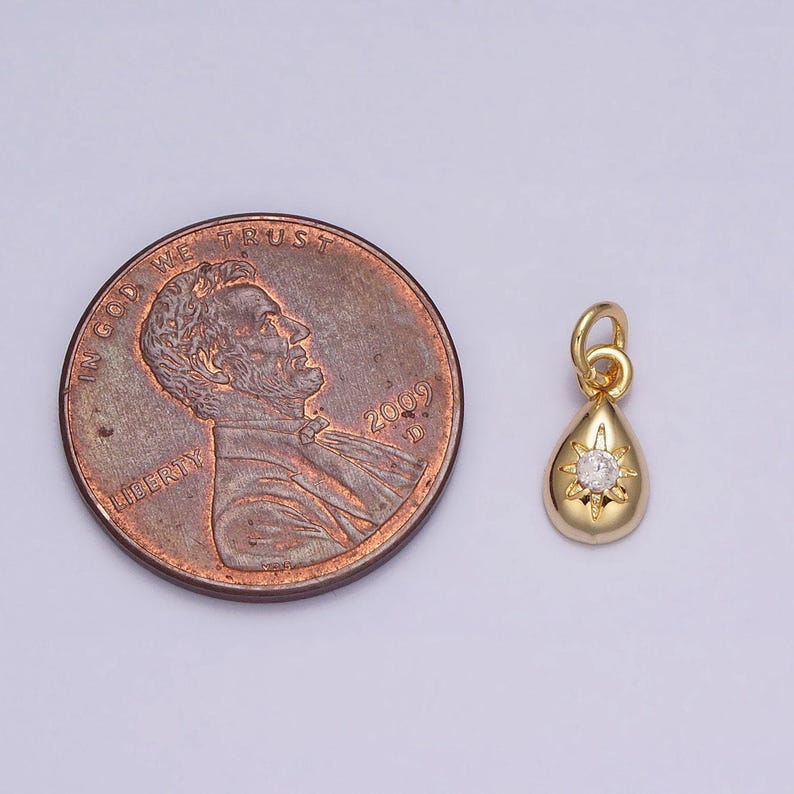 May include: A gold teardrop pendant with a small clear rhinestone in the center. The pendant is shown next to a US penny for size comparison.