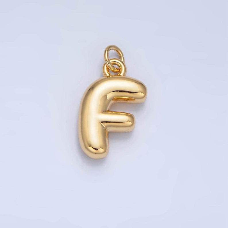 May include: A gold-colored charm in the shape of the letter F. The charm is puffy and has a loop at the top for attaching to a necklace or bracelet.