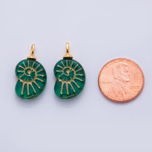 May include: Two green glass pendant charms in the shape of a nautilus shell with gold-colored accents. Each charm has a gold-colored loop at the top. A penny is next to the charms for size comparison.