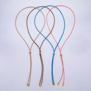 Puede incluir: Cuatro collares ajustables en color beige, marrón, azul y coral. Cada collar presenta un cordón fino con un acento metálico dorado y extremos de cordón a juego. Los collares se muestran sobre un fondo blanco.