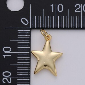 Puede incluir: Un dije de estrella en tono dorado con una superficie lisa y reflectante, que incluye una peque&ntilde;a argolla en la parte superior. El dije est&aacute; colocado junto a una regla negra con marcas blancas, mostrando una anchura de aproximadamente 2 cm.