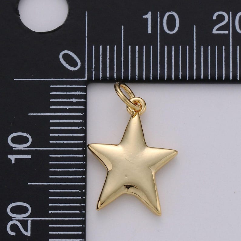 Puede incluir: Un dije de estrella en tono dorado con una superficie lisa y reflectante, que incluye una peque&ntilde;a argolla en la parte superior. El dije est&aacute; colocado junto a una regla negra con marcas blancas, mostrando una anchura de aproximadamente 2 cm.