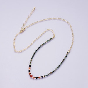 May include: A gold-colored necklace with a beaded design. The necklace features a variety of small, colorful beads, including red, black, and green. The necklace has a gold chain and clasp. The necklace is displayed on a light purple background.