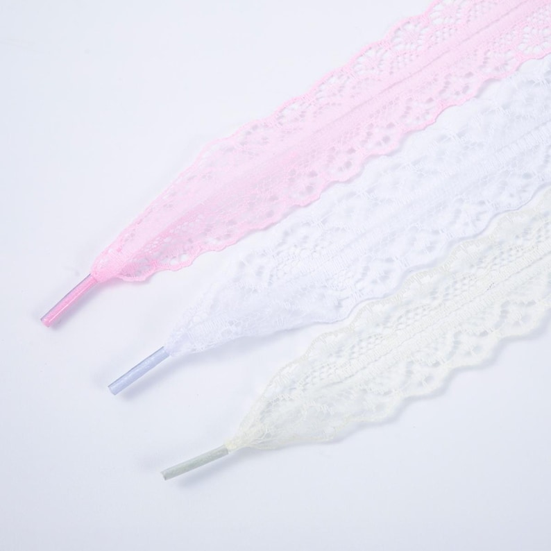 May include: Three decorative shoelaces in pastel colors: pink, white, and pale yellow. Each lace features a delicate, scalloped lace design. The aglets are a matching color to the laces.