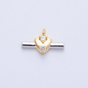 May include: A gold-colored heart-shaped pendant with two clear, round gemstones. The heart is centered on a silver bar. A small loop is at the top for a chain. The pendant is likely for a necklace or bracelet.