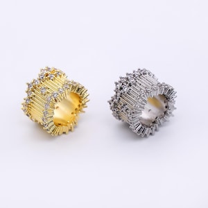 May include: Two rings, one gold-toned and one silver-toned, are displayed against a white background. Both rings feature a cylindrical design with vertical, linear embellishments and small, clear gemstones. The gold ring has a warm, bright finish, while the silver ring has a cool, metallic sheen.