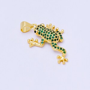 May include: A gold-colored frog pendant with green gemstone accents. The frog is detailed with small green stones covering its body and legs, with black eyes. The pendant has a loop for attaching to a chain or necklace.