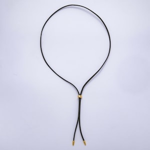 K&ouml;nnte beinhalten: Schwarze Halskette mit goldfarbenem Akzent und goldfarbenen Spitzen. Die Halskette hat ein schlichtes, elegantes Design, wobei eine d&uuml;nne schwarze Kordel eine Schlaufe bildet. Der goldfarbene Akzent ist ein kleines, zylindrisches St&uuml;ck, das entlang der Kordel gleitet.
