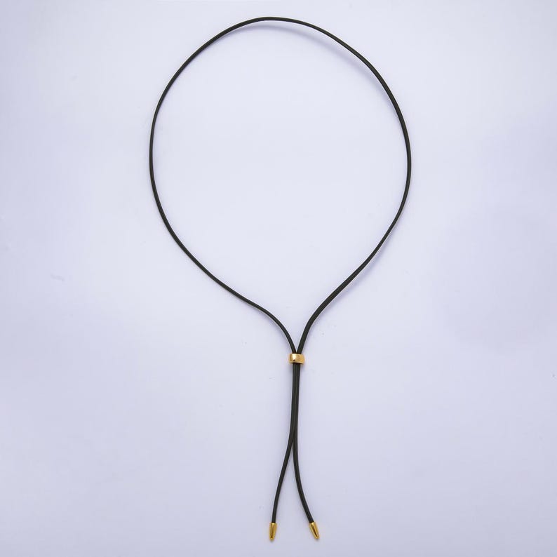 K&ouml;nnte beinhalten: Schwarze Halskette mit goldfarbenem Akzent und goldfarbenen Spitzen. Die Halskette hat ein schlichtes, elegantes Design, wobei eine d&uuml;nne schwarze Kordel eine Schlaufe bildet. Der goldfarbene Akzent ist ein kleines, zylindrisches St&uuml;ck, das entlang der Kordel gleitet.