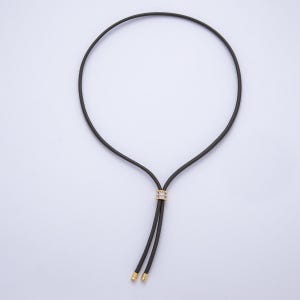 May include: A black cord necklace with a gold-colored accent piece adorned with small, clear stones. The necklace has two dangling ends with gold-colored caps. The design is simple and elegant.