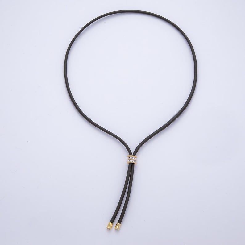 May include: A black cord necklace with a gold-colored accent piece adorned with small, clear stones. The necklace has two dangling ends with gold-colored caps. The design is simple and elegant.