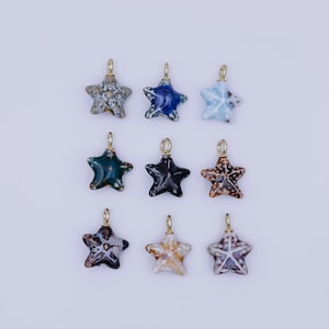 Puede incluir: Nueve colgantes de cerámica con forma de estrella de mar en varios colores, incluyendo azul, verde, marrón y blanco. Cada estrella de mar tiene un lazo dorado para sujetar a un collar o pulsera. Los colgantes están dispuestos sobre un fondo blanco.