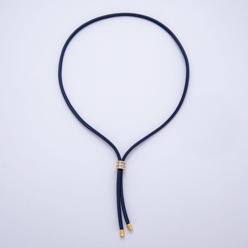 May include: A navy blue necklace with a gold-colored accent. The necklace has a circular design with a gold-colored ring at the center and gold-colored tips at the end of the dangling cords. The necklace is displayed on a white background.