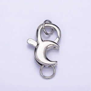 May include: Silver-toned metal lobster clasp with a crescent moon design. The clasp has a small ring on the top and a larger ring on the bottom.