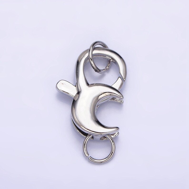May include: Silver-toned metal lobster clasp with a crescent moon design. The clasp has a small ring on the top and a larger ring on the bottom.