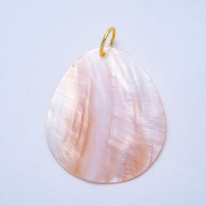 May include: A pearlescent, teardrop-shaped pendant with a gold-colored loop. The pendant has a natural, iridescent surface with shades of pink, white, and cream. The gold loop is attached at the top, ready for a chain or cord.