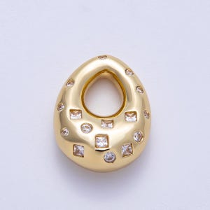 May include: A gold-toned teardrop-shaped pendant featuring a smooth, polished surface embedded with a variety of clear, sparkling stones in both round and square cuts. The pendant has a central open aperture and a reflective, metallic finish.
