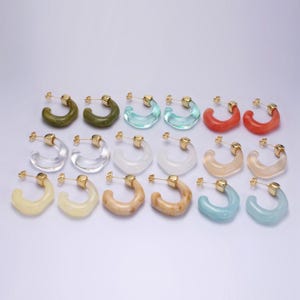 May include: A collection of hoop earrings in various colors, including green, blue, red, and clear. Each earring has a gold-colored post. The earrings are arranged in rows, showcasing their diverse hues and designs.