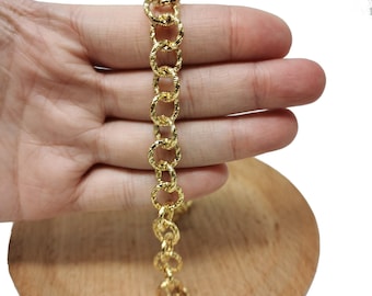 May include: A gold-colored bracelet with a textured, linked chain design. The bracelet is held against a wooden surface, showcasing its intricate details and warm metallic hue. The bracelet is approximately 7 inches long.