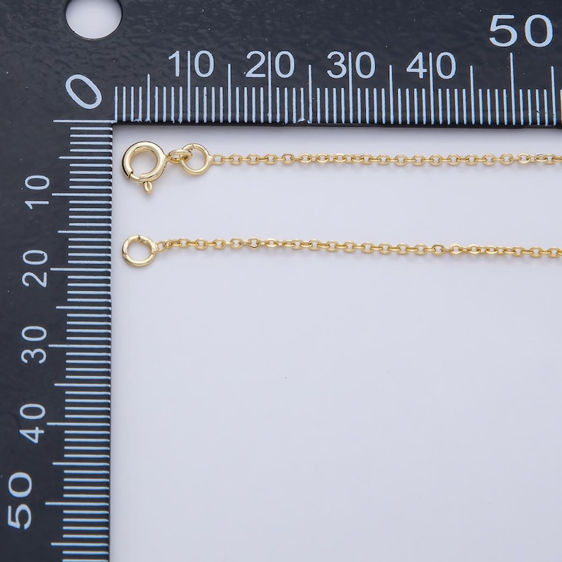K&ouml;nnte beinhalten: Eine zarte goldfarbene Halskette mit Karabinerverschluss und einer kleinen kreisf&ouml;rmigen Schlaufe. Die Kette wird an einem schwarzen Lineal mit wei&szlig;en Markierungen gemessen. Die Halskette ist etwa 25 cm lang.