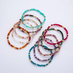 Puede incluir: Una colección de pulseras de cuentas en varios colores, incluyendo turquesa, rojo, verde y morado. Las pulseras presentan pequeñas cuentas redondas y detalles dorados. Las pulseras están dispuestas sobre una superficie blanca.