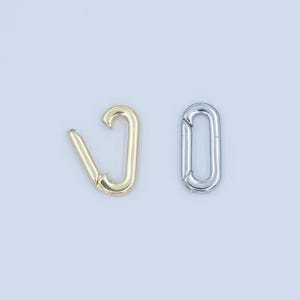 Puede incluir: Dos cierres metálicos, uno dorado y otro plateado, sobre un fondo blanco. El cierre dorado es de tipo mosquetón con resorte, mientras que el plateado es un eslabón ovalado. Ambos son nuevos y sin usar.