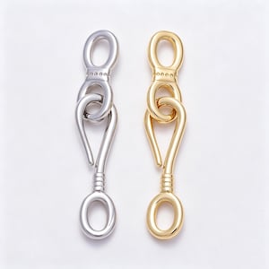 May include: Two metal jewelry clasps, one silver-toned and one gold-toned, each featuring a hook mechanism with a circular loop at the top and bottom. The clasps have a smooth, polished finish with a series of small decorative indentations.