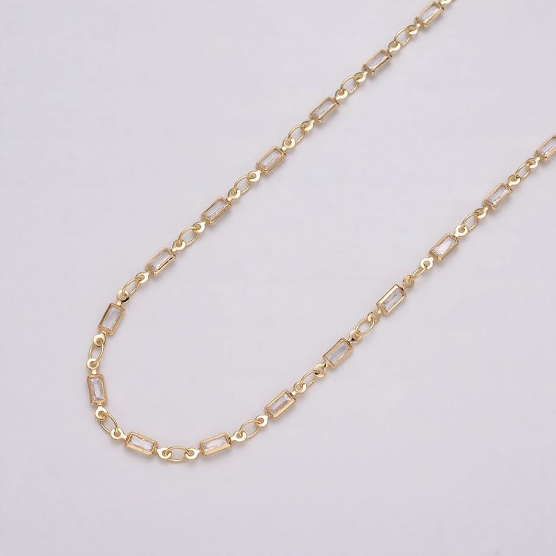 May include: A delicate gold-colored necklace with rectangular, clear crystal accents. The chain is composed of small, interconnected links, creating a refined and elegant accessory. The necklace is displayed against a plain, light background, highlighting its design.