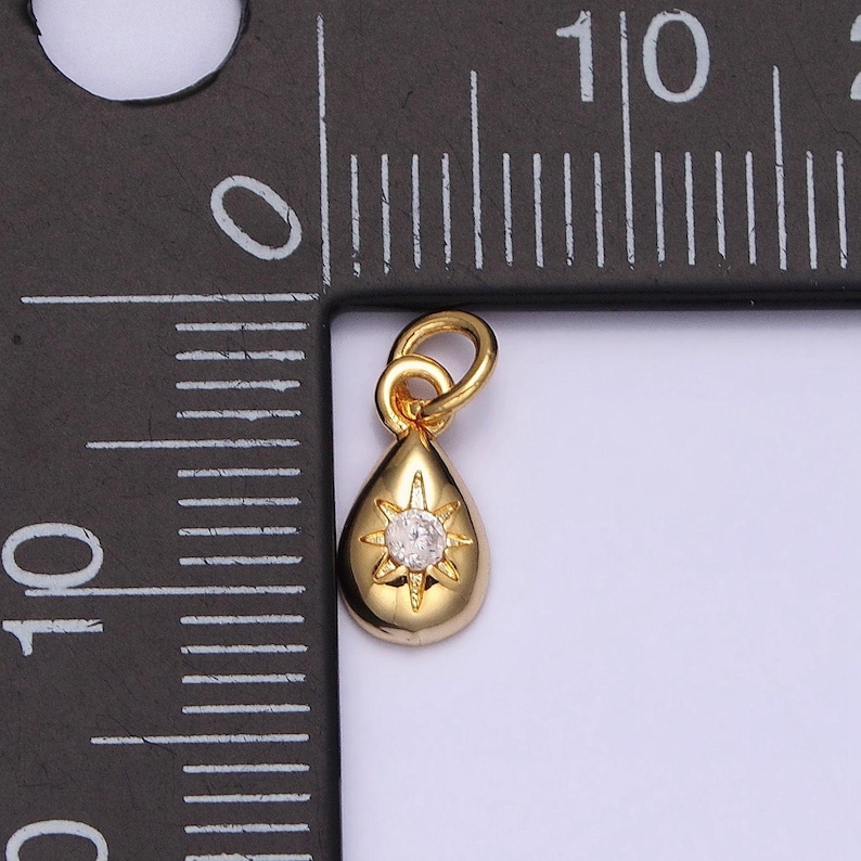 May include: A gold-tone teardrop pendant charm with a clear rhinestone in the center. The pendant has a starburst design around the rhinestone.