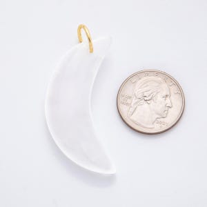 Op de afbeelding: Een witte, halvemaanvormige hanger met een goudkleurige lus om op te hangen. De hanger staat naast een Amerikaanse kwart dollar munt ter vergelijking van de grootte. De hanger is waarschijnlijk gemaakt van een doorschijnend materiaal.
