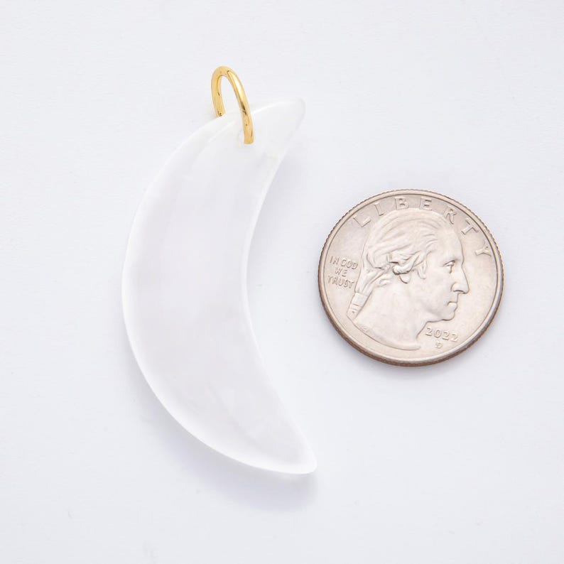 Op de afbeelding: Een witte, halvemaanvormige hanger met een goudkleurige lus om op te hangen. De hanger staat naast een Amerikaanse kwart dollar munt ter vergelijking van de grootte. De hanger is waarschijnlijk gemaakt van een doorschijnend materiaal.