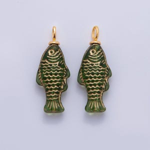 Puede incluir: Dos colgantes verdes con forma de pez y detalles dorados. Los peces tienen escamas y aletas grabadas, y un pequeño lazo dorado en la parte superior para colgar. Estos colgantes son probablemente para la creación de joyas.