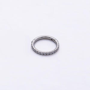 May include: A circular hinged hoop ring made of polished silver-toned metal. The front half of the hoop is embedded with a continuous row of small, clear, round-cut gemstones that catch the light, creating a sparkling, uniform appearance.