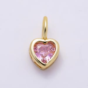 May include: A gold-toned heart-shaped pendant featuring a faceted light pink gemstone set in the center. The pendant includes a small, smooth gold-toned bail at the top for attachment to a chain, showcasing a polished metallic finish throughout.