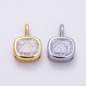 May include: Two square-shaped pendants featuring a central clear faceted gemstone set in a metal frame. One pendant has a polished gold-tone finish, while the other has a polished silver-tone finish. Each piece includes a small vertical bail at the top.