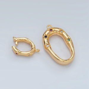 Könnte beinhalten: Zwei goldfarbene Metall-Schmuckverbinder. Einer ist eine einfache ovale Schlaufe, der andere ist ein Oval mit einer strukturierten Oberfläche und bunten Edelsteinen.