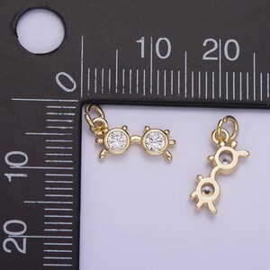 May include: Two gold-tone charm pendants in the shape of eyeglasses with clear rhinestone accents. The charms are shown next to a ruler for size reference.