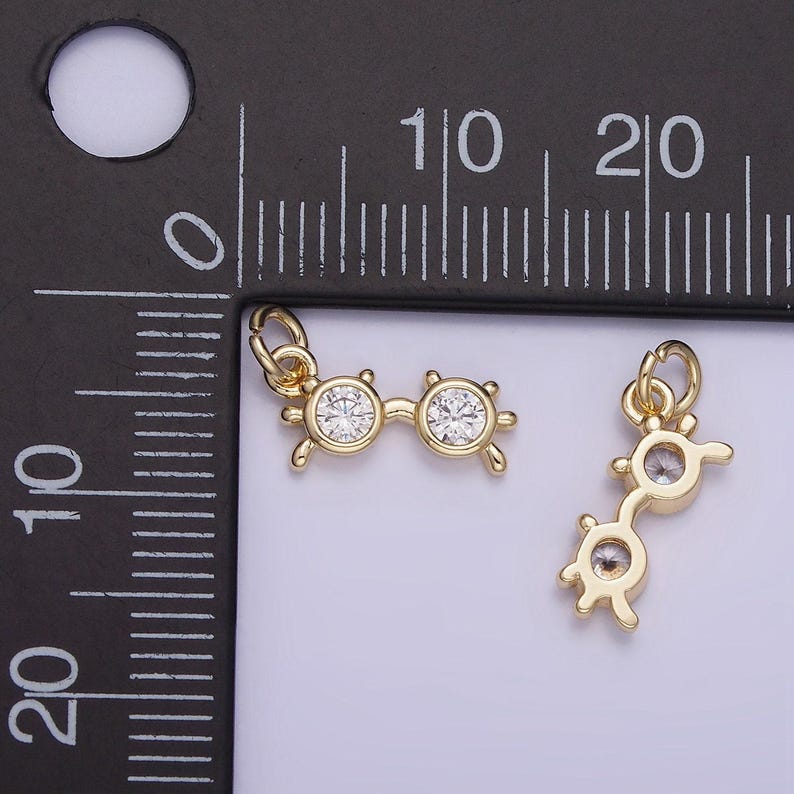 May include: Two gold-tone charm pendants in the shape of eyeglasses with clear rhinestone accents. The charms are shown next to a ruler for size reference.