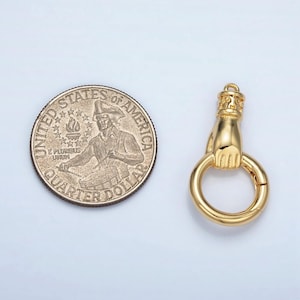 K&ouml;nnte beinhalten: Ein goldfarbener Anh&auml;nger mit einer Hand, die einen Ring h&auml;lt, neben einem US-Vierteldollar. Der Anh&auml;nger hat ein detailliertes Design einer Hand und einer dekorativen Oberseite. Der Vierteldollar hat Standardgr&ouml;&szlig;e.