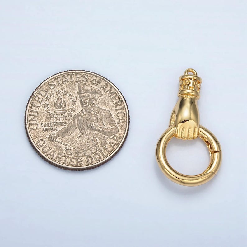 K&ouml;nnte beinhalten: Ein goldfarbener Anh&auml;nger mit einer Hand, die einen Ring h&auml;lt, neben einem US-Vierteldollar. Der Anh&auml;nger hat ein detailliertes Design einer Hand und einer dekorativen Oberseite. Der Vierteldollar hat Standardgr&ouml;&szlig;e.