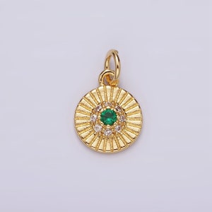 May include: A gold-colored pendant with a circular design. The pendant features a central green gemstone surrounded by smaller clear gemstones. The outer edge has a sunburst pattern. A small loop is at the top for a chain.