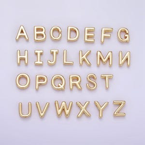 May include: A set of gold-coloured alphabet letter charms, arranged in rows. Each letter is in a bold, rounded font, and the letters are evenly spaced. The letters are displayed against a light purple background.